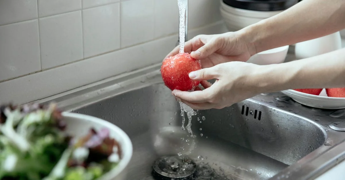 Mains lavant une pomme rouge fraîche sous l'eau courante dans un évier de cuisine.