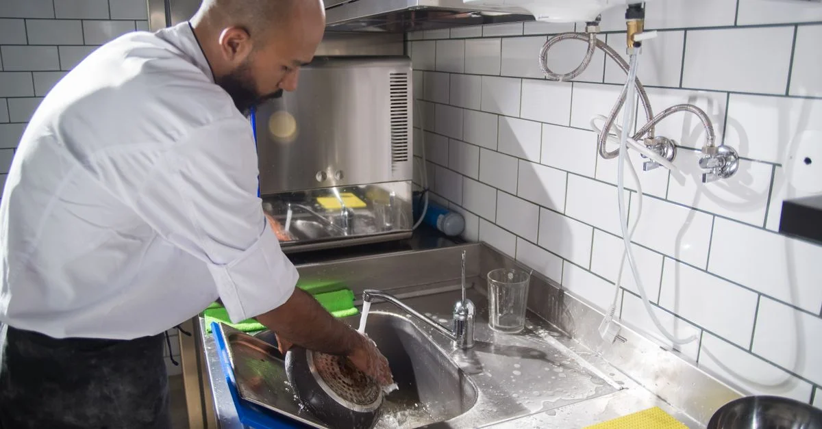 Un chef nettoie des ustensiles de cuisine dans un évier en acier inoxydable, maintenant des normes d'hygiène.