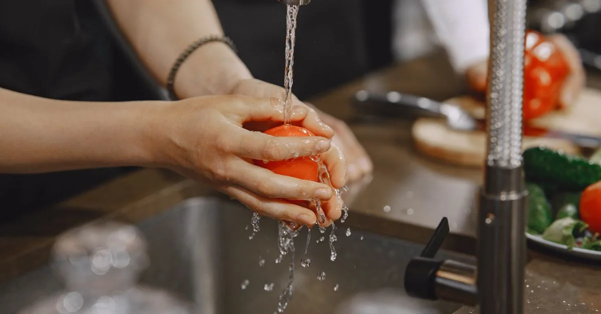 Gros plan sur des mains lavant une tomate rouge mûre sous un robinet de cuisine. L'eau éclabousse en action.