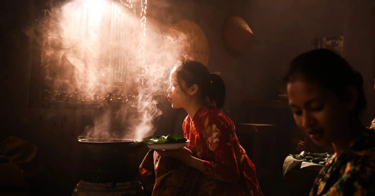 Deux femmes en tenue traditionnelle cuisinent dans une pièce doucement éclairée avec de la vapeur chaude qui s'élève.