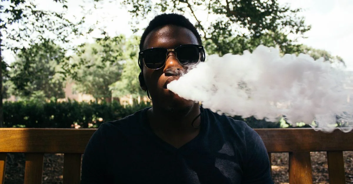 Un jeune homme portant des lunettes de soleil expire de la vapeur en s'asseyant sur un banc en bois dans un cadre extérieur ensoleillé.