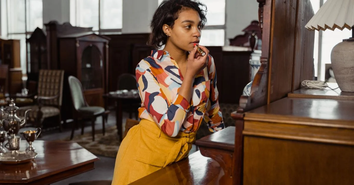 Femme élégante appliquant du rouge à lèvres tout en se tenant dans un magasin de meubles vintage avec des meubles et une décoration en bois.