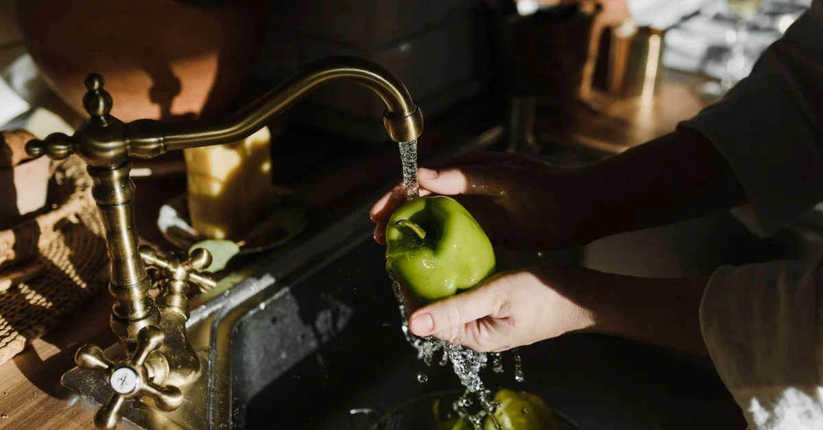 Des mains lavant un poivron vert frais sous un robinet vintage dans un cadre de cuisine chaleureux et rustique.