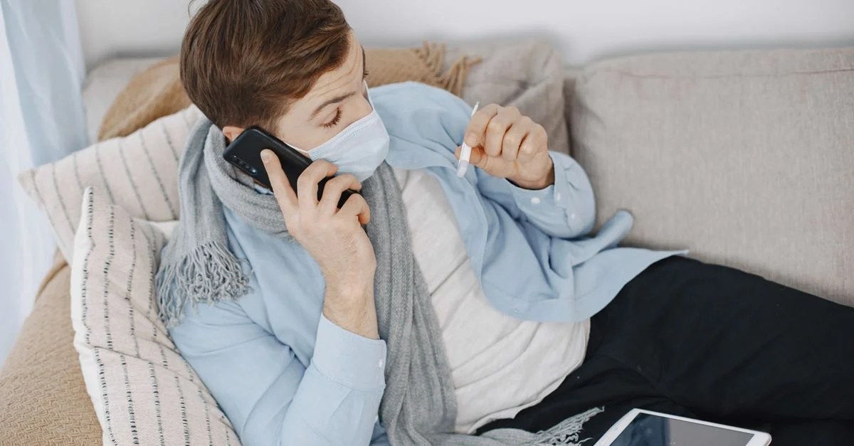 Homme utilisant des services de télémédecine tout en étant malade à la maison. Allongé sur le canapé avec un téléphone et un thermomètre.