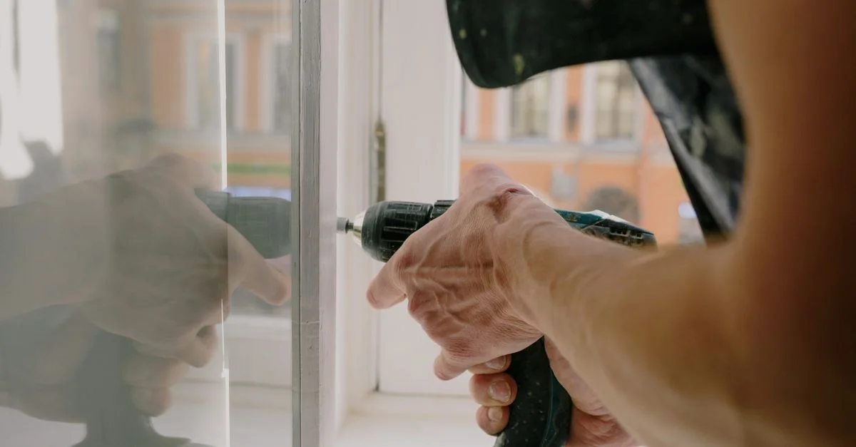 Gros plan des mains d'un bricoleur utilisant une perceuse pour fixer une fenêtre dans un appartement.