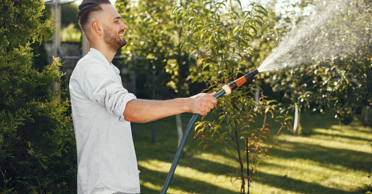 Homme souriant en chemise blanche arrosant des plantes dans un jardin verdoyant. Journée lumineuse et claire.
