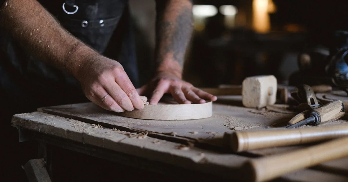 Gros plan sur les mains d'un artisan ponçant soigneusement un disque en bois dans un atelier faiblement éclairé.