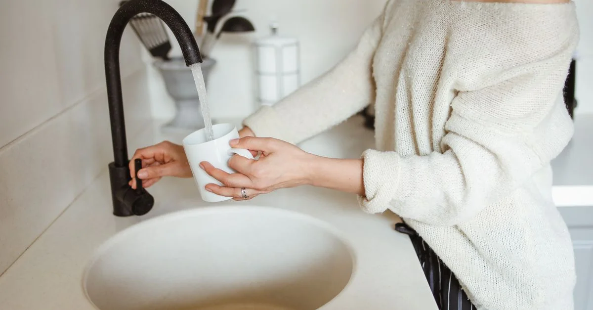 Une femme remplissant une tasse blanche d'eau provenant d'un robinet noir dans une cuisine moderne.